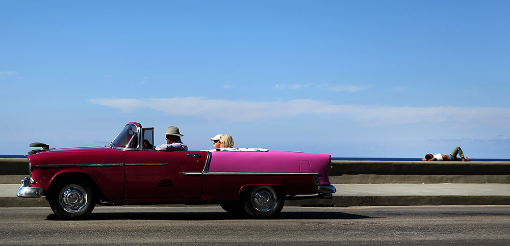 Paraíso tropical. Una pareja de turistas disfruta un viaje a bordo de un auto clásico que funciona como taxi en la ciudad de La Habana, Cuba, mientras dejan atrás a un hombre que toma el sol en pleno malecón.